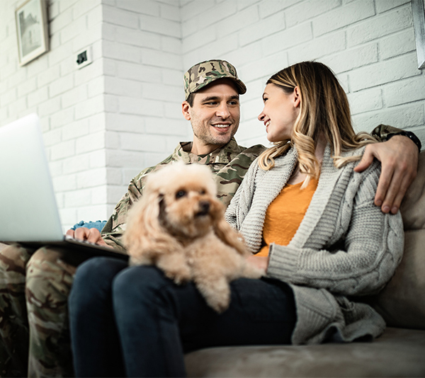 Happy military couple sitting on couch with small dog and a laptop.