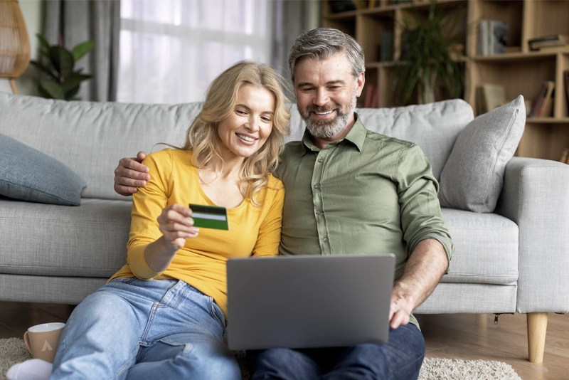 Happy couple sitting on living room floor with laptop and credit card