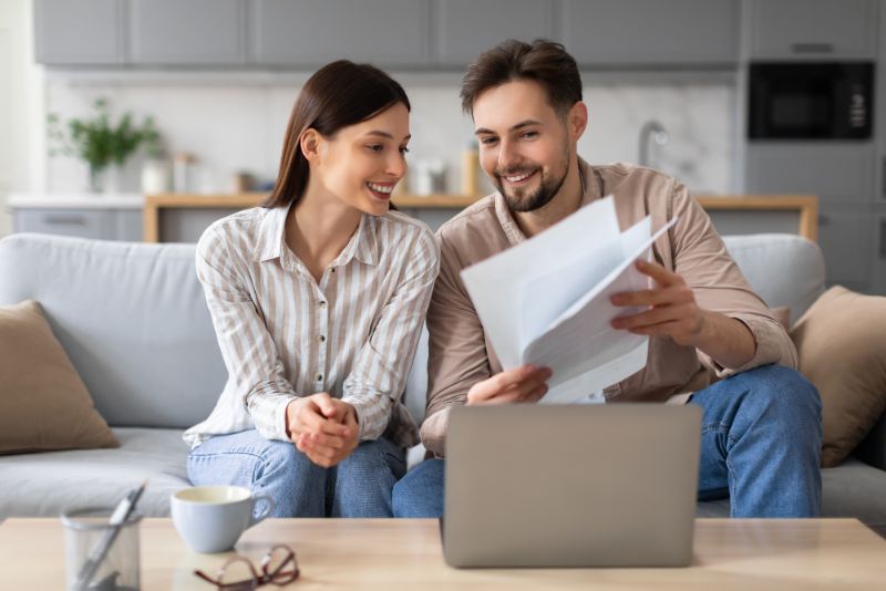 Smiling couple sitting on a soft reviewing documents in front of a laptop